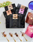 A beautifully arranged flat lay featuring various packets of Bestsellers of Magic Tea Starter Set- 60-75 cup sampler box in black, brown, and pink packaging. Also included are a blue glass, spoons with loose leaf tea and rose petals, and vibrant flowers on a white background, capturing the essence of Magic Hour.