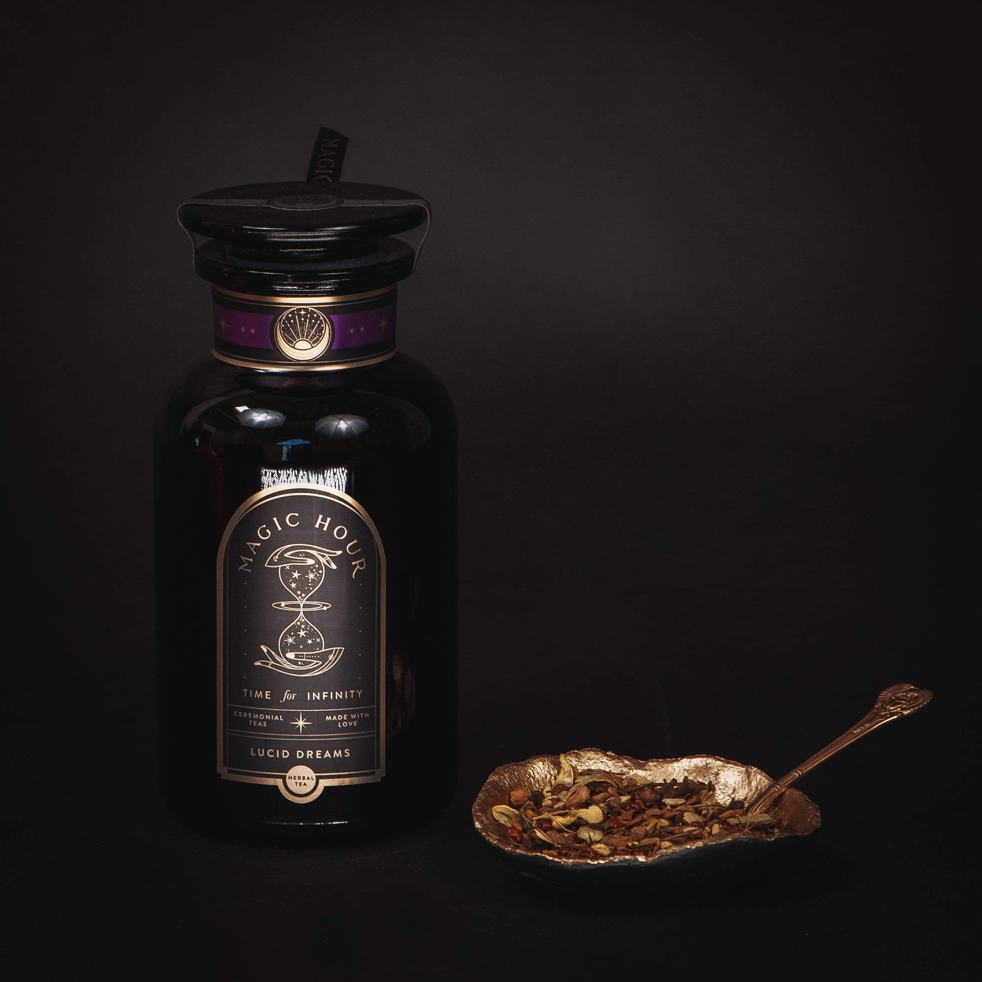 A black glass bottle labeled "Magic Hour - Best Sellers of Magic Hour Box" with a gold accent and a black cap sits next to a decorative, gold-colored leaf-shaped dish holding a spoon and an array of dried herbs and spices, evoking the essence of organic loose leaf tea against a dark background.