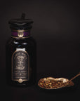 A black glass bottle labeled "Magic Hour - Best Sellers of Magic Hour Box" with a gold accent and a black cap sits next to a decorative, gold-colored leaf-shaped dish holding a spoon and an array of dried herbs and spices, evoking the essence of organic loose leaf tea against a dark background.