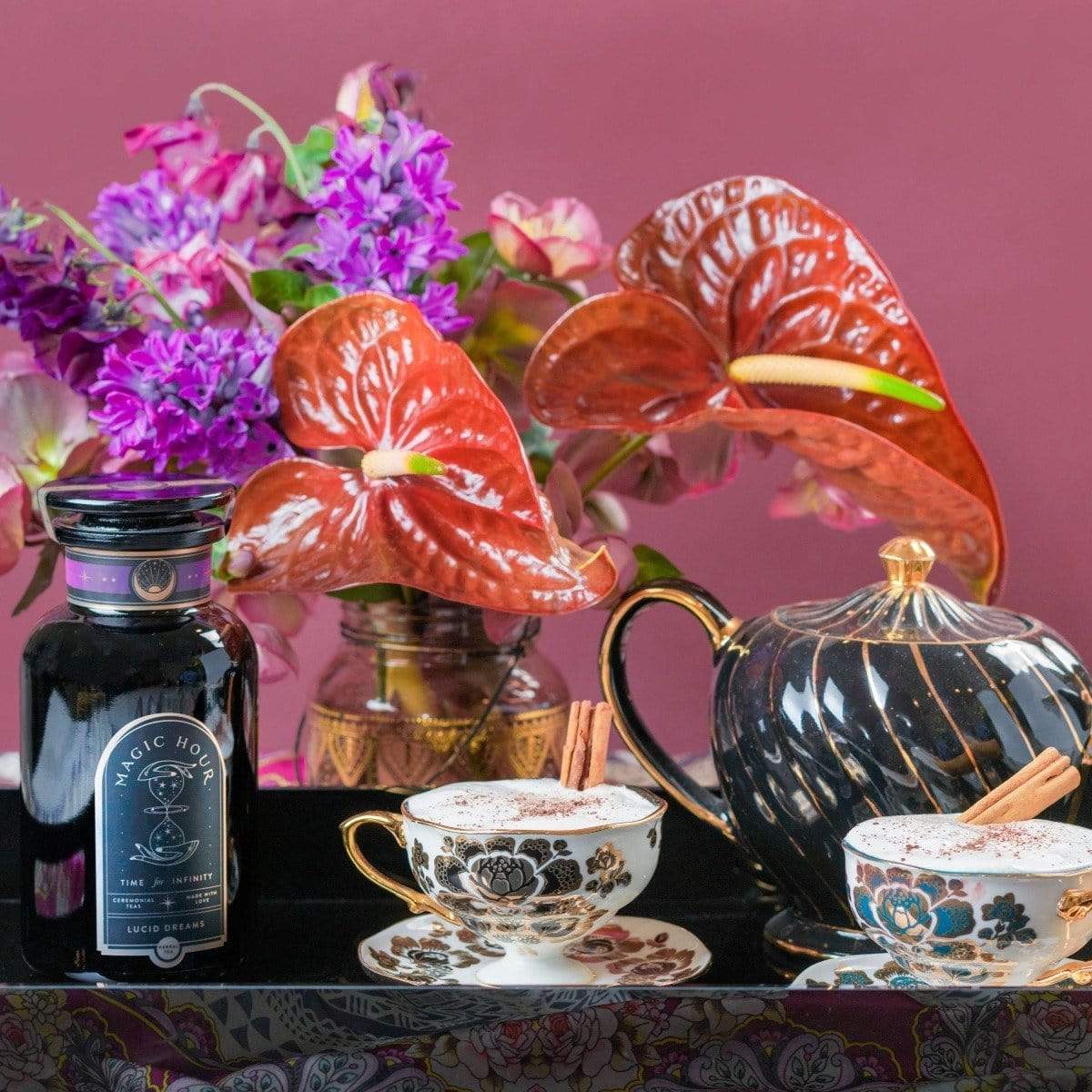 A vibrant floral arrangement featuring purple and red flowers stands behind a black teapot, two patterned teacups with cinnamon sticks, and a jar labeled "Drift into Sleep Ritual Gift" by Magic Hour on a tray. The background is a rich, warm pink.