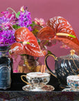 A vibrant floral arrangement featuring purple and red flowers stands behind a black teapot, two patterned teacups with cinnamon sticks, and a jar labeled "Drift into Sleep Ritual Gift" by Magic Hour on a tray. The background is a rich, warm pink.