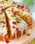 A close-up of a slice of cake topped with glossy white icing, bits of pistachios, and rose petals. The cake sits on a decorative peach-colored plate, with the rest of the cake visible in the background, also topped with icing and garnishes. Enjoy this delightful treat with a cup of Libra- Pistachio-Rose Persian Love Cake Tea with White Pearls & Shatavari by Magic Hour.