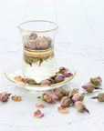 A clear glass cup filled with Magic Hour's Libra- Pistachio-Rose Persian Love Cake Tea with White Pearls & Shatavari, featuring dried rosebuds, sits on a matching glass saucer with a gold rim. The cup and saucer rest on a white lace doily. Dried rosebuds are scattered around the saucer on the doily.