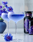 Two elegant cocktail glasses filled with a vibrant blue drink sit on a white surface, reminiscent of the soothing Sapphire - Apple-Plum-Spice White Tea by Magic Hour. A purple flower is placed beside one glass. In the background, there is a vase with similar purple flowers and two dark bottles adorned with intricate purple designs.