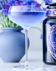 A delicate cocktail glass filled with lavender-colored Sapphire - Apple-Plum-Spice White Tea from Magic Hour is in the foreground. Behind it, there's a blue vase containing purple flowers and a stylish dark bottle with a geometric design on the label, all set against a light background.