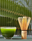 A jar of Magic Hour Kansai-Kyoto Ceremonial Matcha, a bamboo whisk, a small bowl of green matcha tea, and a bamboo scoop are arranged in front of a textured glass panel with green fern leaves visible in the background. The setup is aesthetic and minimalist, highlighting the finest organic tea products.
