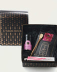 A gift box with "Magic Hour" written on it, containing various items including a small jar with a pink label, a gold spoon, a small glass bottle, a packet of Sip Wellness: Ritual Kit for Immunity & Joy Tea, and a roll of Magic Hour brand incense. The box interior has black shredded paper for padding.
