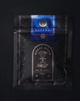 A black package of "Moon Lover: Herbal Immunitea Ritual Gift" organic tea with celestial designs and gold detailing from Magic Hour. The label features a blue section with a tree logo and text, while the main section showcases an hourglass graphic accompanied by "Time for Infinity" and the loose leaf tea type description.
