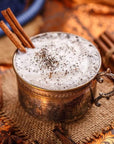 A vintage metal cup filled with frothy white beverage topped with ground spices and garnished with cinnamon sticks. It's placed on a textured mat surrounded by loose leaf tea, cinnamon sticks, and star anise, set against a warm, rustic background. The scene features Magic Hour's Queen of the South: Delicious Cocoa Detox Tea.