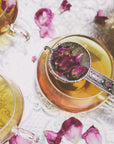 A close-up of three tea cups filled with light brown Wild Indian Roses by Magic Hour, placed on an ornate silver tray. Rose petals are scattered around and one silver strainer filled with rose petals rests on top of one cup. The scene has a delicate, elegant feel, embodying the essence of fine Organic Tea.
