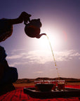 A person is pouring Sin Eraser™ : Puerh Tea from Magic Hour from a teapot into glasses on a tray set on a red cloth in a desert. The silhouetted figure is against a setting sun, creating a serene atmosphere with the sky transitioning from light to dark.