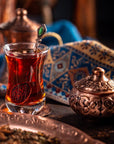 A glass of Sin Eraser™: Puerh Tea by Magic Hour with a decorative metal spoon sits on a copper saucer. The background features an intricate patterned cloth and an ornate copper container with a lid. The scene is richly colored and exudes a warm, traditional atmosphere.