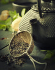 A close-up of a textured black teapot beside a tea strainer filled with loose Gamma-Aminobutyric Acid (GABA) Oolong Tea by Magic Hour. In the background, blurred elements include mint leaves and slices of ginger on a rustic wooden surface. The overall lighting is soft and warm, creating a cozy atmosphere perfect for calm nourishment.