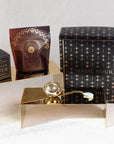 A collection of organic tea products is elegantly displayed on metallic risers. The set includes a packet of loose leaf tea, a beautifully crafted Sip Royaltea Gift Set box, a tea strainer shaped like a disco ball with a chain, and a large black box with "Magic Hour" text adorned with gold stars against the white background.