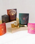A group of four colorful tea packaging pouches with intricate designs displayed on gold platforms. There is also a gold tea infuser and a black Magic Hour box on the left side. The background is a plain, light-colored fabric, highlighting the elegance of The Queen's Sampler Set by Magic Hour.
