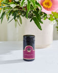 A jar labeled "Raspberry Earl White Traveler" white tea from Magic Hour sits on a light gray surface. The jar has a dark top and a maroon label with gold accents, highlighting its organic origins. In the background, there is a white vase filled with a colorful floral arrangement, including pink, orange, and white blossoms and greenery.