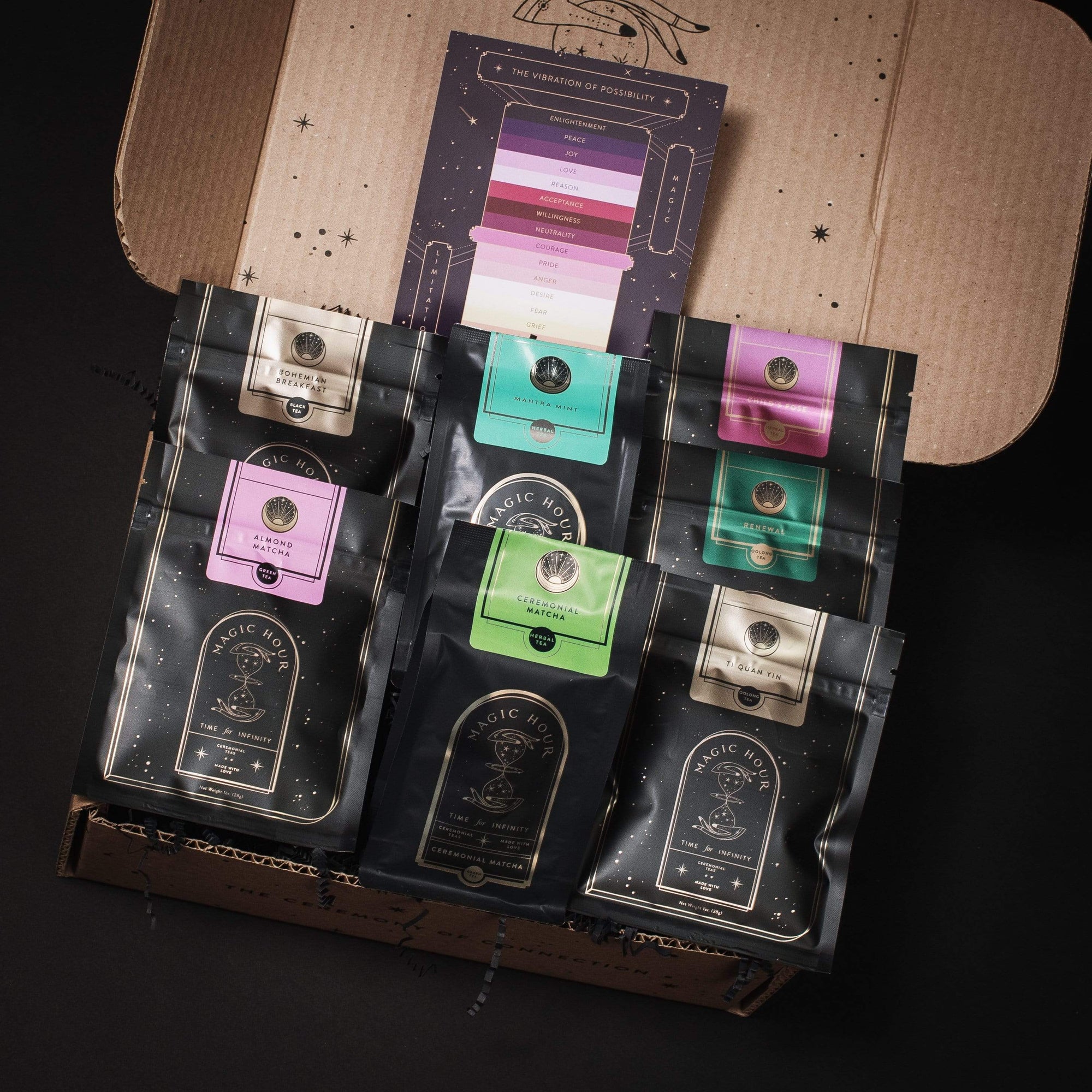 A cardboard box with the lid open reveals seven black packages of coffee with various colored labels arranged neatly inside. Behind the packages, there's a booklet titled "The Persuasion of Possibility," evoking the enchanting allure akin to sampling Club Magic Hour's Self-Guided 14 Day Ceremonial Sensory Tea Cleanse Kits.