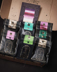 A cardboard box with the lid open reveals seven black packages of coffee with various colored labels arranged neatly inside. Behind the packages, there's a booklet titled "The Persuasion of Possibility," evoking the enchanting allure akin to sampling Club Magic Hour's Self-Guided 14 Day Ceremonial Sensory Tea Cleanse Kits.