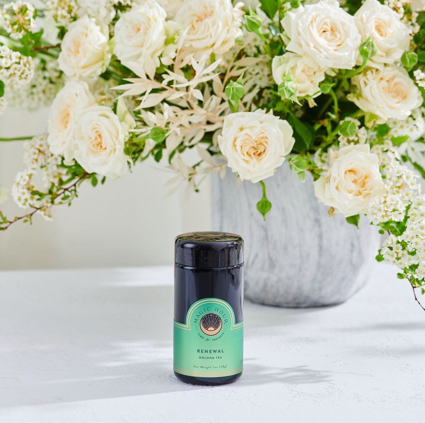 A jar of "Renewal: Peach-Goji-Rose Oolong Tea Traveler" by Magic Hour sits on a white surface in front of a vase filled with cream-colored roses and greenery. The jar is dark with a green label and black lid, showcasing the premium loose leaf tea inside.