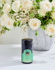 A jar of "Renewal: Peach-Goji-Rose Oolong Tea Traveler" by Magic Hour sits on a white surface in front of a vase filled with cream-colored roses and greenery. The jar is dark with a green label and black lid, showcasing the premium loose leaf tea inside.