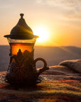 A decorative glass teacup with an ornate metal handle and lid sits on a colorful woven blanket. The cup contains Rise & Shine Black Tea by Club Magic Hour, illuminated by the warm glow of a Magic Hour Tea sunset in the background, casting a serene and tranquil ambiance.
