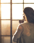 A person with long hair is standing indoors, holding a mug of Rise & Shine Black Tea from Club Magic Hour, and looking out of a large window at a bright morning sun. They are dressed in a cozy sweater, and the atmosphere is warm with a soft, golden light filtering through the window.