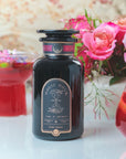 A dark glass bottle labeled "Club Magic Hour: Ruby Moon™ : Hibiscus Elderberry Tea" stands on a table. Next to it are two glasses containing red liquids with garnishes. In the background, a vase filled with vibrant, pink flowers adds a pop of color to the scene.