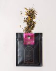 A black and gold Magic Hour tea packet labeled "Symbeeosis: Beautifying Immunitea for the Queen Bee" lies flat on a white surface. The packet is opened with loose leaf tea and flower petals spilling out onto the surface above the packet, offering a glimpse of this exquisite organic tea blend.