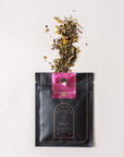 A black resealable bag labeled "Sip Wellness: Ritual Kit for Immunity & Joy" by Magic Hour with a pink sticker on top is shown, spilling a mix of organic loose-leaf tea onto a light surface. Various dried ingredients, including flowers and herbs, are visible in the tea blend.