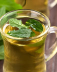 A clear glass cup filled with hot Organic Spearmint Herbal Tisane garnished with fresh mint leaves. Another glass with the same beverage is blurred in the background. The setting includes some more organic mint leaves on a wooden surface beside the cups.