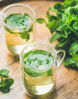 Two clear glass mugs filled with refreshing Organic Spearmint Herbal Tisane are placed on a wooden surface. Fresh mint leaves, including aromatic spearmint, are visible inside the mugs and scattered nearby. A large bunch of organic mint is situated to the right, adding a touch of green to the scene.