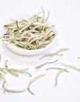 A small white bowl filled with Silver Moon White Tea leaves from Club Magic Hour is placed on a clean, white surface. Several thin, elongated leaves are scattered around the bowl, creating a minimalist and clean visual. The pale color of the Silver Moon White Tea adds to the serene aesthetic.