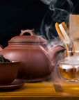 A steaming clay teapot and bowl with loose Organic Lapsang Souchong: Smoky Black Tea leaves are placed on a wooden surface. Two clear teacups filled with the rich, smoky-flavored tea and a set of wooden utensils are in the background, against a dark backdrop. Steam rises from both the teapot and cups, creating a cozy ambiance.