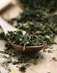 A wooden spoon is filled with loose, dried tea leaves. More dried tea leaves are scattered on the wooden surface around the spoon, suggesting a rustic and natural setting. The organic tea leaves are likely an herbal or green variety, reminiscent of a serene Magic Hour Mao Jian (Emerald) Green Tea of Good Fortune moment.