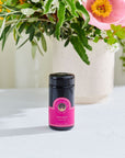 A small, dark glass jar with a pink label reading "Soulmate: Chocolate-Raspberry-Rose Black Tea Traveler by Magic Hour" sits on a white surface. In the background, a white ceramic pot showcases a vibrant floral arrangement with green foliage and colorful flowers, creating an enchanting backdrop fit for sipping Organic Tea.