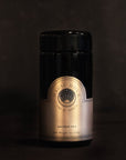 A dark container of Magic Hour ceremonial tea, known as the Violet Traveler Jar, encased in violet glass with a detailed and elegant label that displays the brand name in a circular, gold-accented design, against a plain, dark background.
