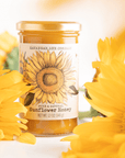 A jar of Savannah Bee Co. Honey - Sunflower is surrounded by vibrant yellow sunflowers. The label features an illustration of a sunflower and contains 12 ounces (340 grams) of pure and natural sunflower honey, sourced from the rich fields of Ukraine and Romania.