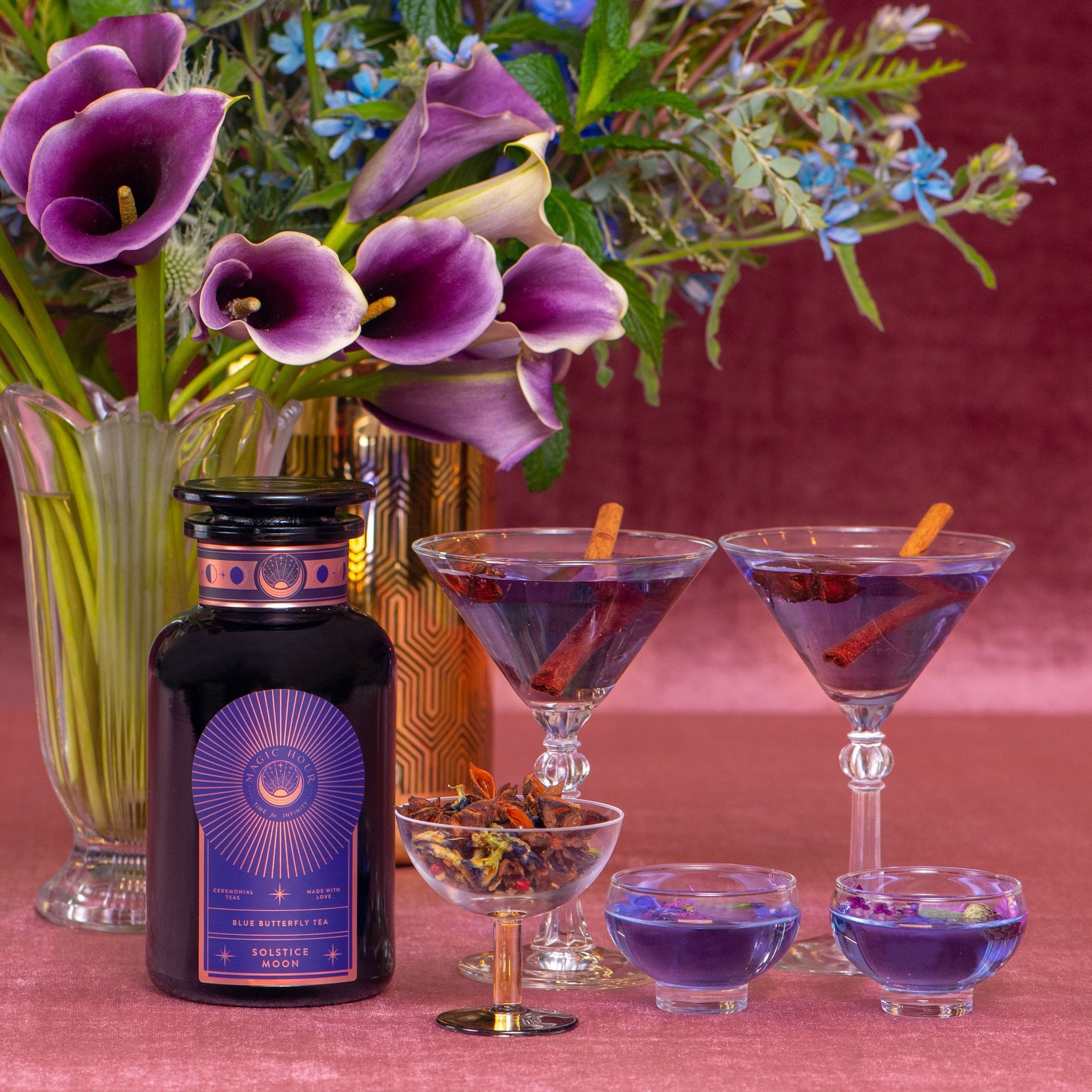 A bottle of Solstice Moon™ gin sits beside a bouquet of flowers. Two martini glasses contain purple cocktails garnished with cinnamon sticks. Nearby, a small glass cup holds dried botanicals, and two small shallow cups hold blue liquid. The background is pink, evoking the magic of Winter Solstice celebrations.