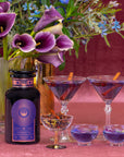 A bottle of Solstice Moon™ gin sits beside a bouquet of flowers. Two martini glasses contain purple cocktails garnished with cinnamon sticks. Nearby, a small glass cup holds dried botanicals, and two small shallow cups hold blue liquid. The background is pink, evoking the magic of Winter Solstice celebrations.