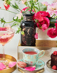 A vibrant arrangement features a floral bouquet in the background and a bottle with an "Astrology Bundle: Aries, Taurus, Gemini - Magic Hour" label at the center. In the foreground, there is a decorative teacup and saucer filled with organic tea, a glass of pink beverage, a chocolate cake topped with berries, and macarons.