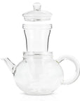 The Hand Blown 32oz Glass Teapot with Infuser is made from heat-resistant borosilicate glass and includes an infusion chamber and lid. Its rounded body features a spout on the left side and a handle on the right, with a removable glass infuser basket sitting inside its top opening. The teapot is currently empty.