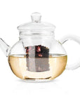 The Hand Blown 32oz Glass Teapot with Infuser has a transparent, round body complete with a handle, spout, and lid. Crafted from heat-resistant borosilicate glass, this teapot includes a removable glass infuser basket. Hibiscus tea leaves steep inside the light yellow liquid, clearly visible through the glass.