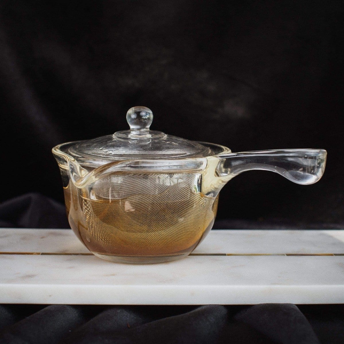 A transparent glass teapot with a matching glass lid is placed on a white marble slab. The teapot, perfect for brewing Zhena's Essential Favorites Tea Collection, features a subtle tint of orange around the base. Set against a dark background, the translucent design and elegant handle are beautifully highlighted.