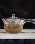 A transparent glass teapot with a matching glass lid is placed on a white marble slab. The teapot, perfect for brewing Zhena's Essential Favorites Tea Collection, features a subtle tint of orange around the base. Set against a dark background, the translucent design and elegant handle are beautifully highlighted.