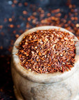 A rustic stone mortar is filled to the brim with finely shredded, reddish-brown, organic Queen of the Harvest : Herbal Relaxation Tea by Magic Hour. The dark background is scattered with additional loose leaf tea, highlighting the vibrant tone and texture of the rooibos.