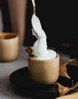 A spoon is lifting a swirl of whipped cream from a small beige cup. The cup sits on a black saucer with a cinnamon stick, dried leaves, and hints of loose leaf tea nearby. Another similar cup is in the background. The setting has a warm and cozy atmosphere, reminiscent of Magic Hour's Queen of the Harvest: Herbal Relaxation Tea moments.