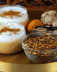 Two glass cups filled with frothy, spiced beverages are on a golden tray, each topped with ground cinnamon. Beside the cups is a bowl of mixed spices and scattered ginger roots. A whole nutmeg and a piece of turmeric complete the scene, perfect for an organic tea experience with Magic Hour's Queen of the Harvest: Herbal Relaxation Tea.