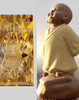 On the left, a shiny gold tea package with intricate designs is showcased, boasting its Caramel Apple Crisp Oolong blend. On the right, a small, smiling statue of a bald child monk in a robe stands on a rock, looking content. The background is light grey. In the center, The Topaz Gemstone Box adds an extra touch of elegance to the display.