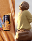 A decorative scene featuring a Buddha tea ceremony statuette on the right, and on the left, The Topaz Gemstone Box with an intricate golden label of Caramel Apple Crisp Oolong tea, a tarot card, and a gold fan against a rich brown backdrop.