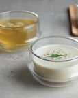 A table displays two glass bowls with handles. The foreground bowl is filled with creamy white soup garnished with herbs and black pepper. The background bowl contains clear broth with visible ingredients. A wooden tray, adorned with spices and a delicate cup of Kinto Double-Walled Teacups, is partially visible.